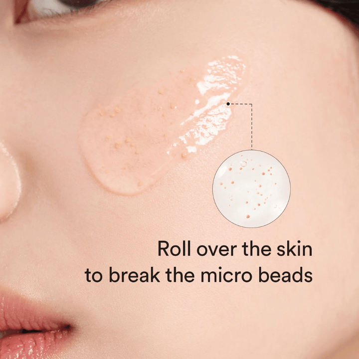 Gros plan sur une joue avec un sérum contenant de petites micro-perles appliqué sur la peau. Un zoom circulaire montre les perles plus clairement. Le texte dit « Rouler sur la peau pour briser les micro-perles ».