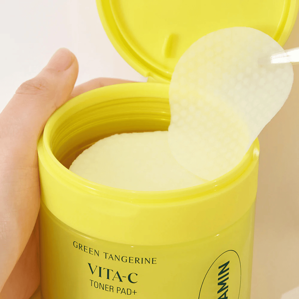 Une main utilise une pince en plastique pour prendre un tampon de toner texturé dans un récipient jaune clair marqué "GREEN TANGERINE VITA-C TONER PAD+" de la marque "gooda