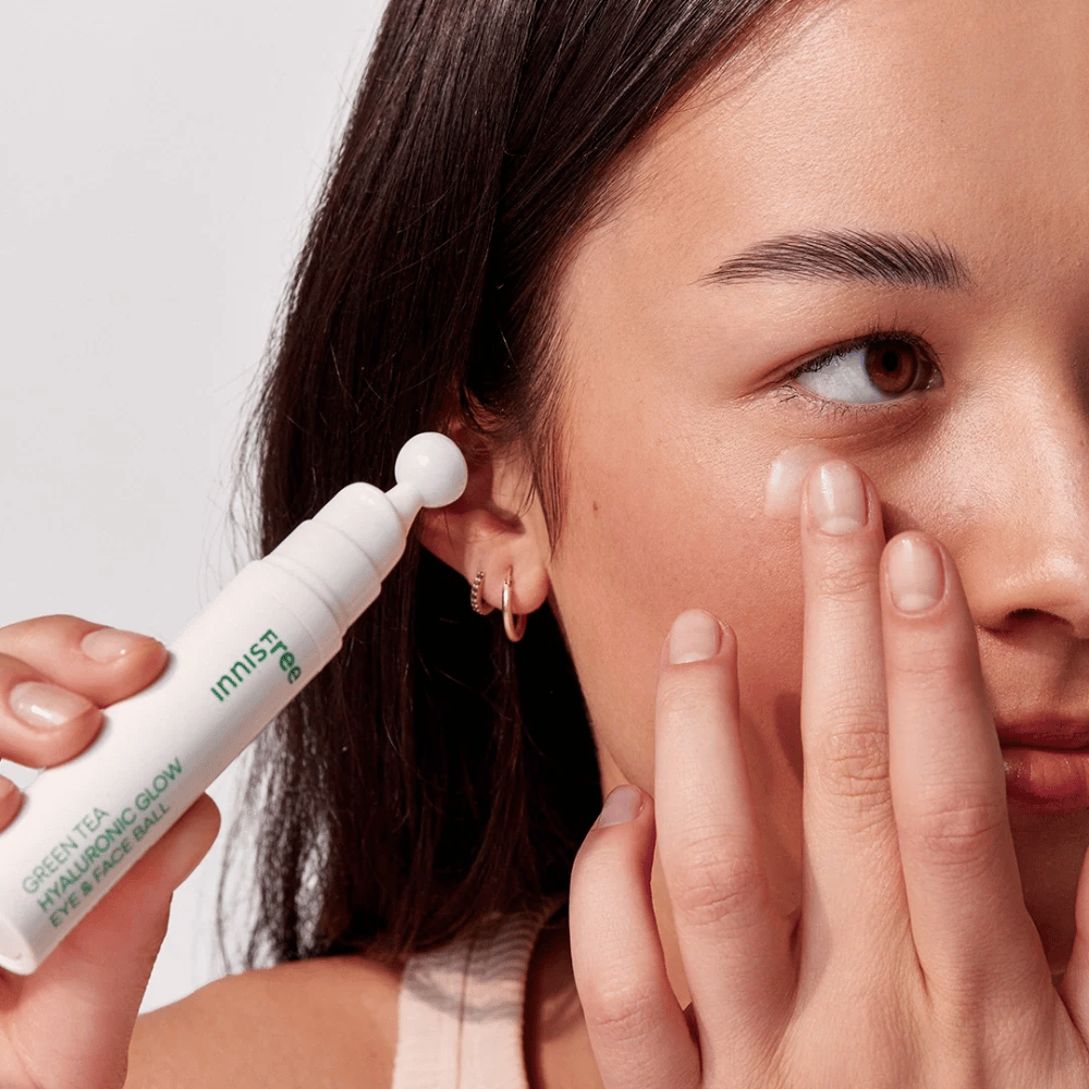 Une personne applique Innisfree Green Tea Hyaluronic Glow Eye & Face Ball sur sa peau sous les yeux. Le produit a une partie applicatrice blanche et ronde et est dans une bouteille blanche avec du texte vert. Le visage de l'utilisateur est partiellement visible et se concentre sur l'application du produit.