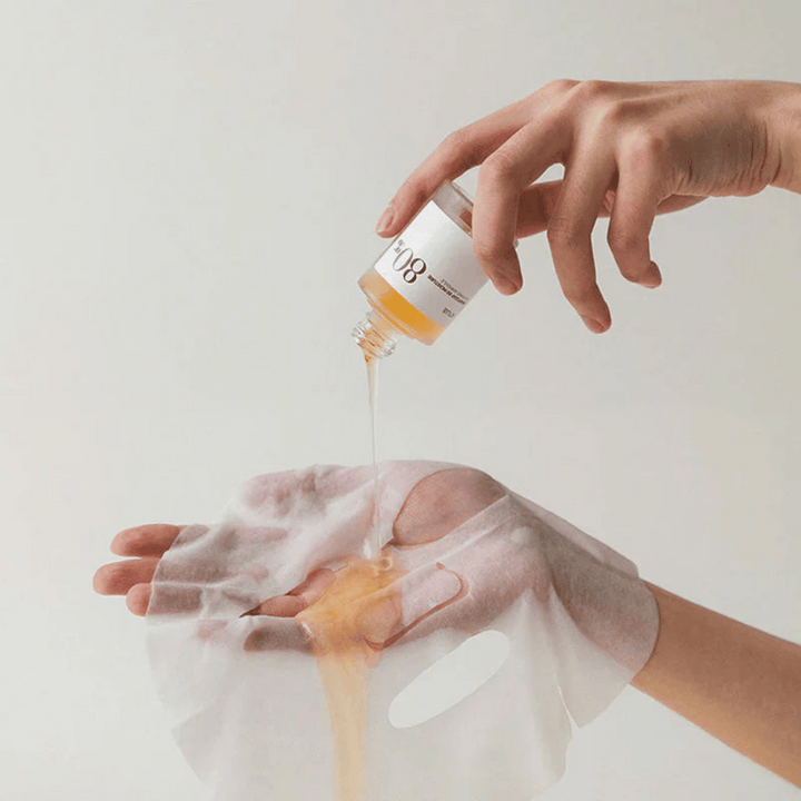 Une main verse un liquide doré et épais d'une petite bouteille marquée '108' sur un masque facial transparent tenu dans une autre main. L'arrière-plan est neutre et uniforme pour mettre en valeur le produit.
