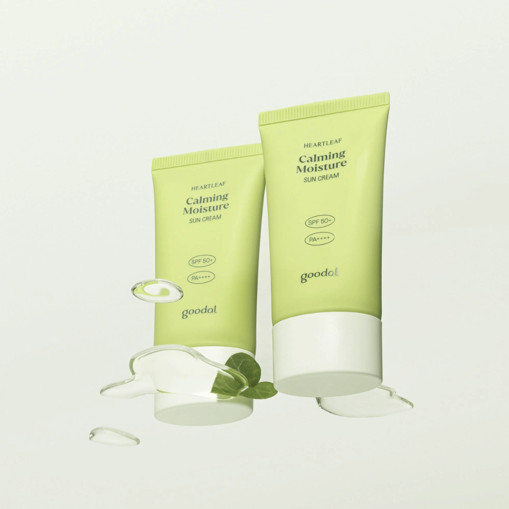 Deux tubes de Goodal Heartleaf Calming Moisture Sun Cream SPF50+ PA++++ entourés de feuilles de plantes et de gouttes de sérum sur un fond clair.