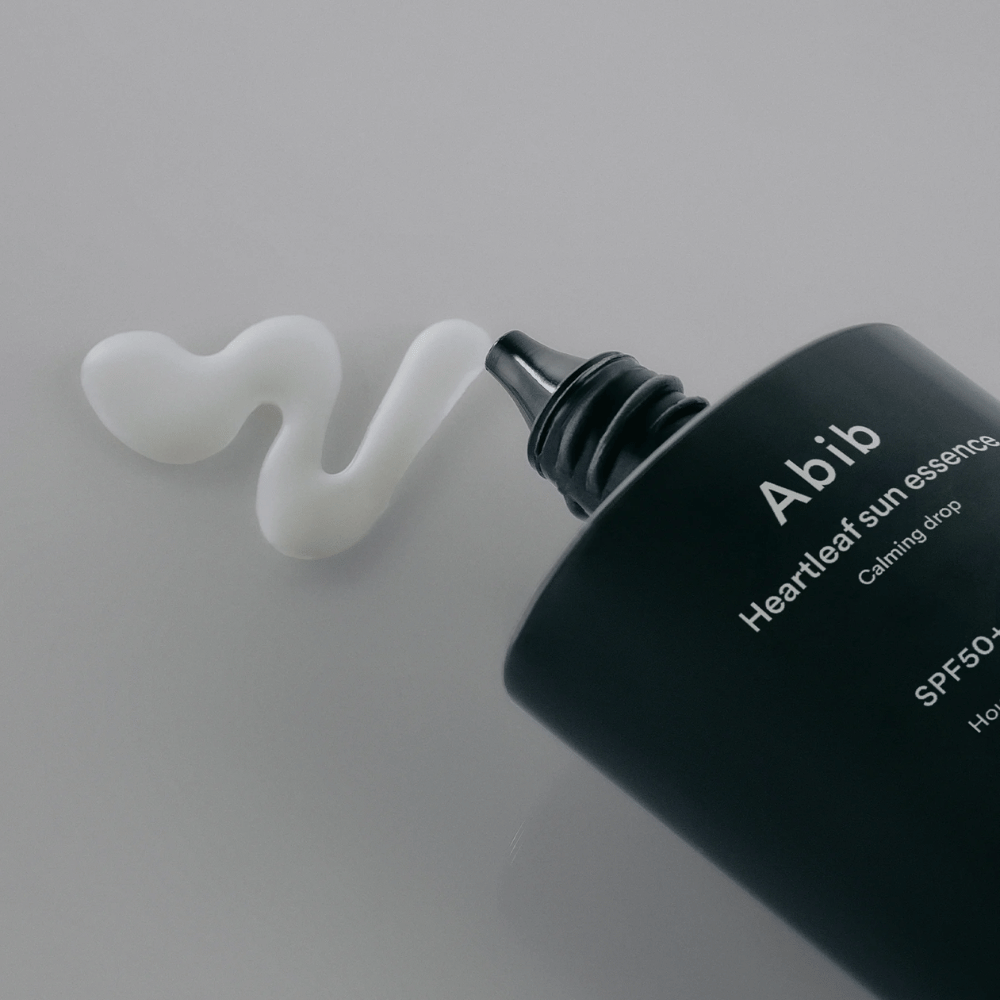 Gros plan sur Abib Heartleaf Sun Essence Calming Drop avec la crème solaire pressée en une ligne ondulée blanche sur une surface claire.