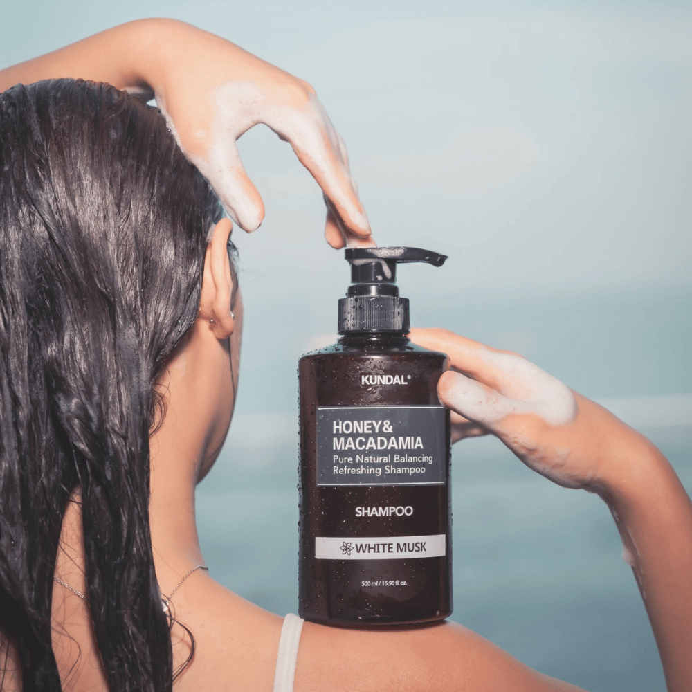 Femme aux cheveux mouillés applique de la mousse et tient KUNDAL Honey & Macadamia Nature Shampoo White Musk sur fond de mer.