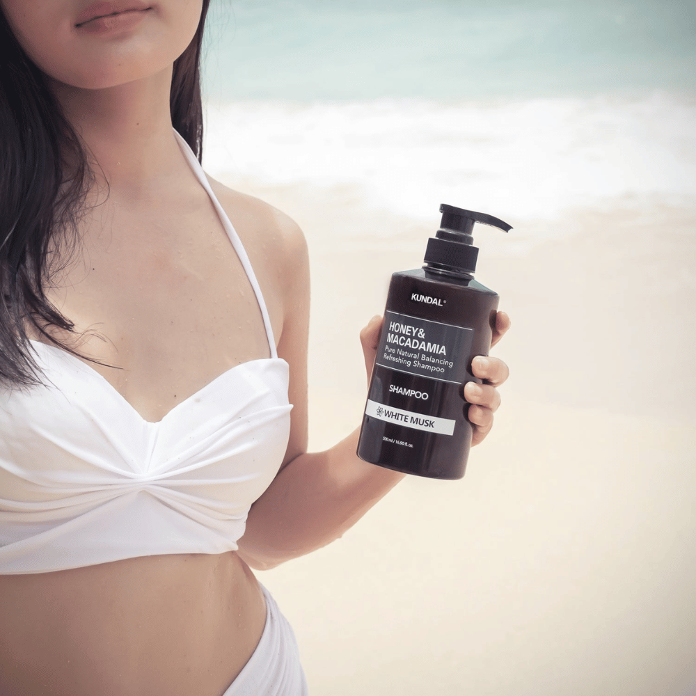 Femme en bikini blanc tient KUNDAL Honey & Macadamia Nature Shampoo White Musk sur une plage avec la mer en arrière-plan.