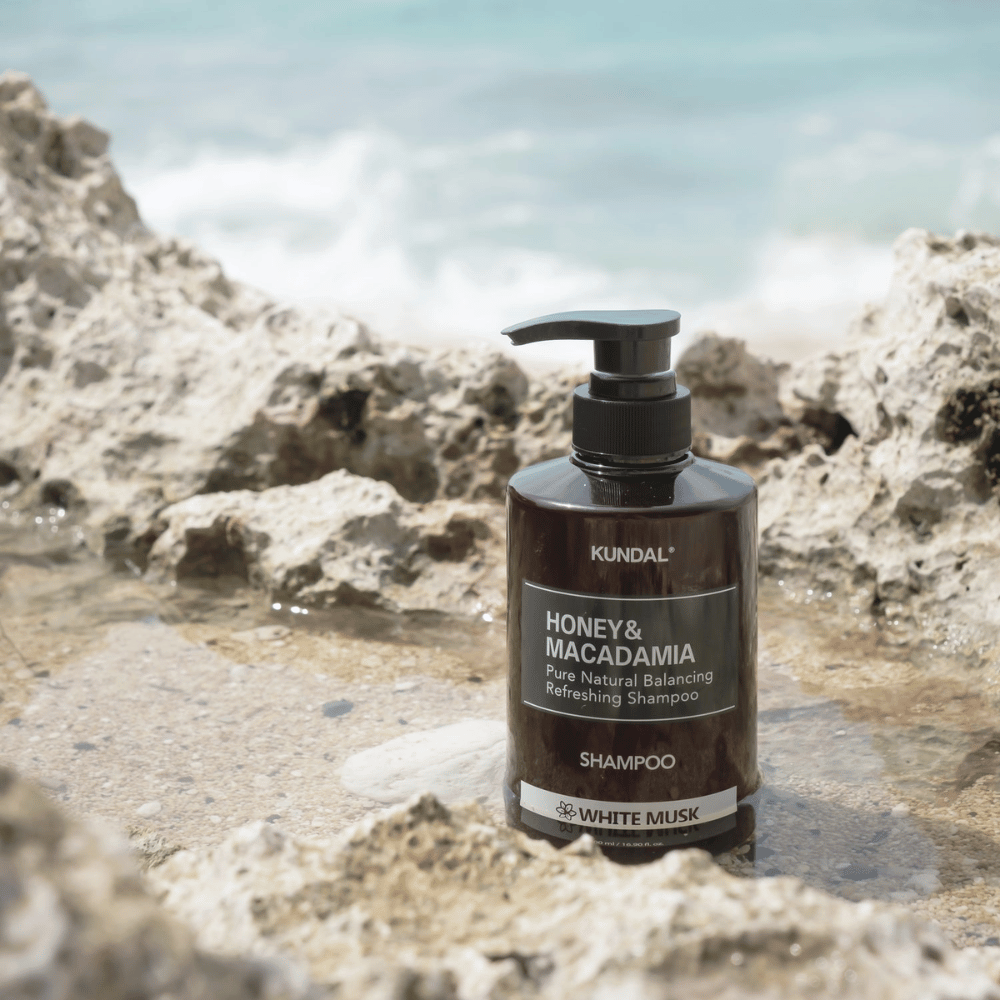 KUNDAL Honey & Macadamia Nature Shampoo White Musk placé sur des rochers au bord de la mer, un produit de soins capillaires naturel avec un parfum frais.