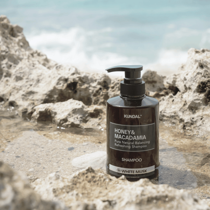 KUNDAL Honey & Macadamia Nature Shampoo White Musk placé sur des rochers au bord de la mer, un produit de soins capillaires naturel avec un parfum frais.