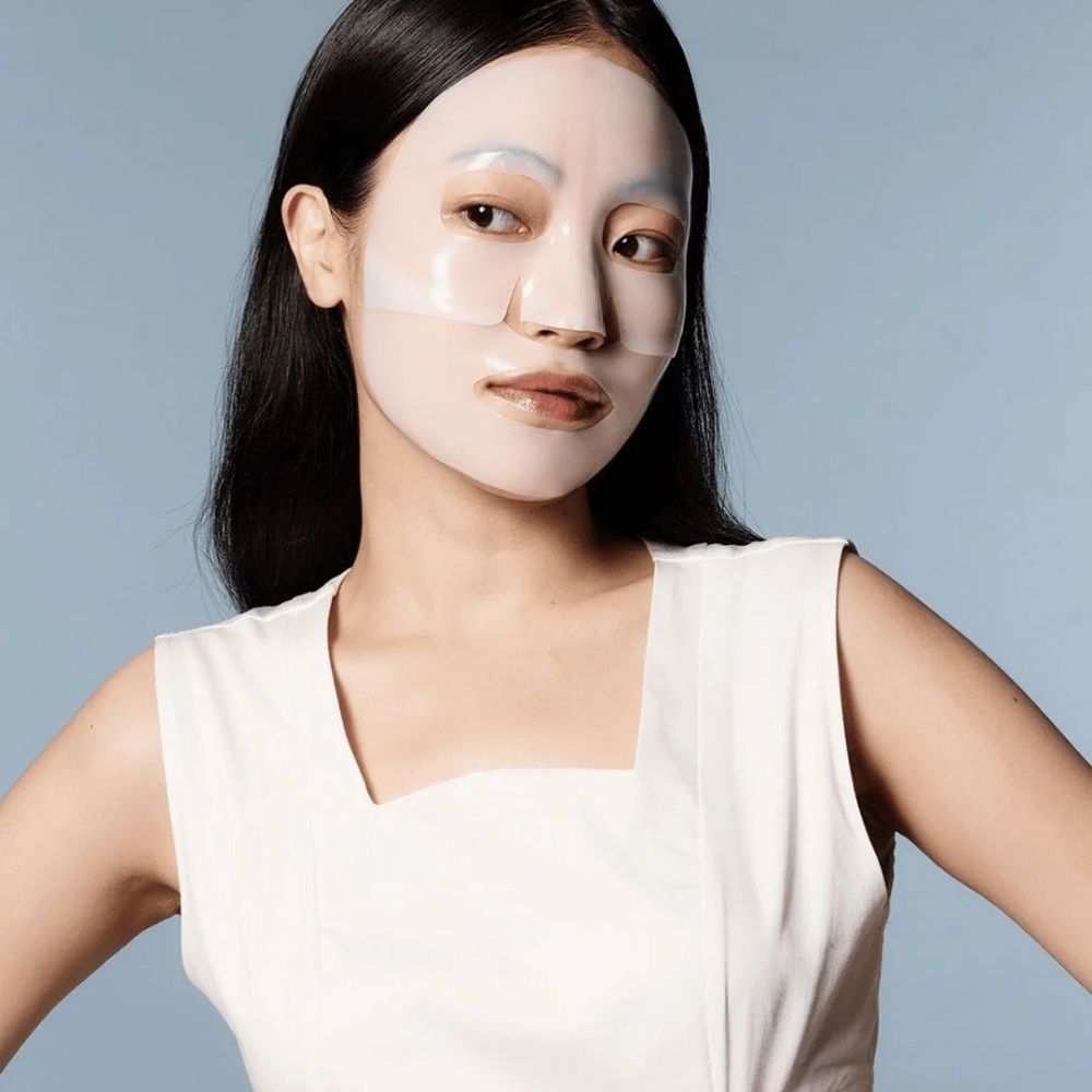 Une femme en robe blanche sans manches porte un masque facial transparent qui couvre presque tout son visage. Le masque donne une impression de soin de la peau avancé et a l'apparence d'un traitement de beauté, conçu pour hydrater en profondeur et traiter la peau.