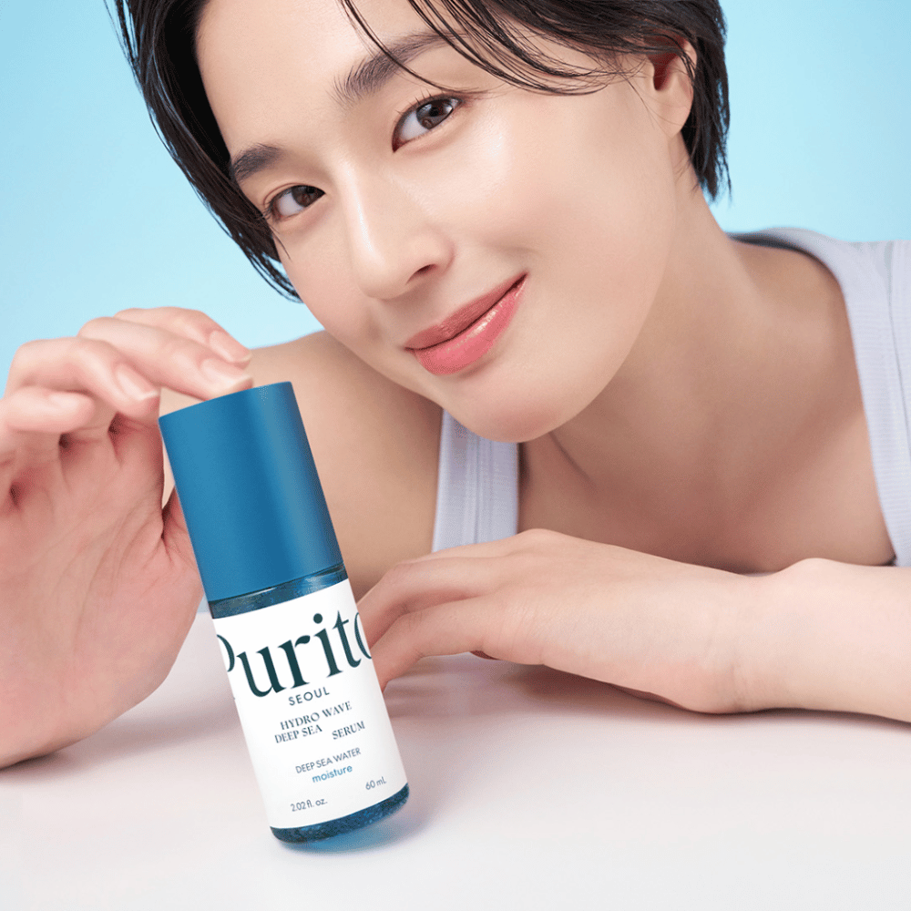 Une femme aux cheveux courts tient un sérum bleu de Purito, Hydro Wave Deep Sea Serum. Sa peau semble radieuse et hydratée, mettant en valeur le produit au centre. L'arrière-plan est bleu clair, ce qui donne une sensation fraîche et propre. Le produit est approfondi dans le soin de la peau.