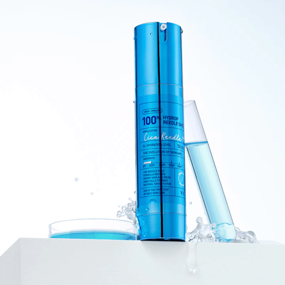 Bouteille bleue VT Cica Hyalon Hydrop Reedle Shot 100hL debout sur une surface blanche avec des éclaboussures d'eau, à côté d'un tube à essai rempli de liquide bleu et d'un bouchon rond avec le même liquide.