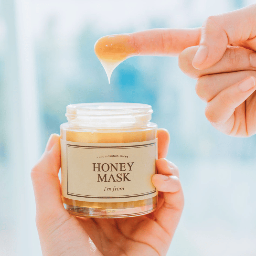 Une main tient un pot ouvert de "I'm from Honey Mask", et un doigt trempe dans le masque doré sur un fond clair.