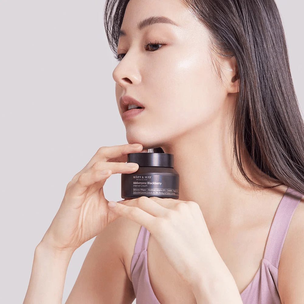 Une femme tient un pot de MARY & MAY Idebenone Blackberry Intense Cream. Elle a un teint éclatant et sourit légèrement, vêtue d'un haut sans manches. Le produit est une crème intensive avec idébénone et complexe de mûre, destinée à la fermeté et à l'hydratation de la peau.