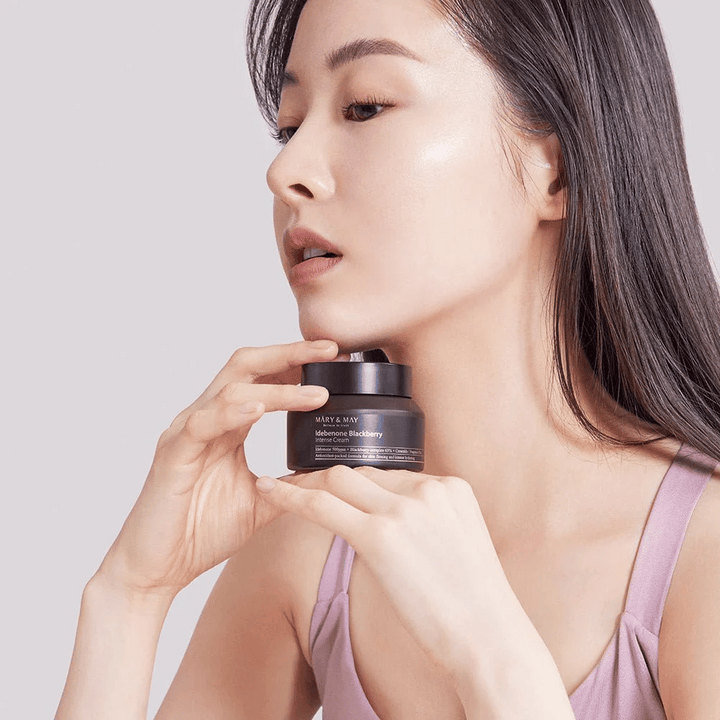 Une femme tient un pot de MARY & MAY Idebenone Blackberry Intense Cream. Elle a un teint éclatant et sourit légèrement, vêtue d'un haut sans manches. Le produit est une crème intensive avec idébénone et complexe de mûre, destinée à la fermeté et à l'hydratation de la peau.