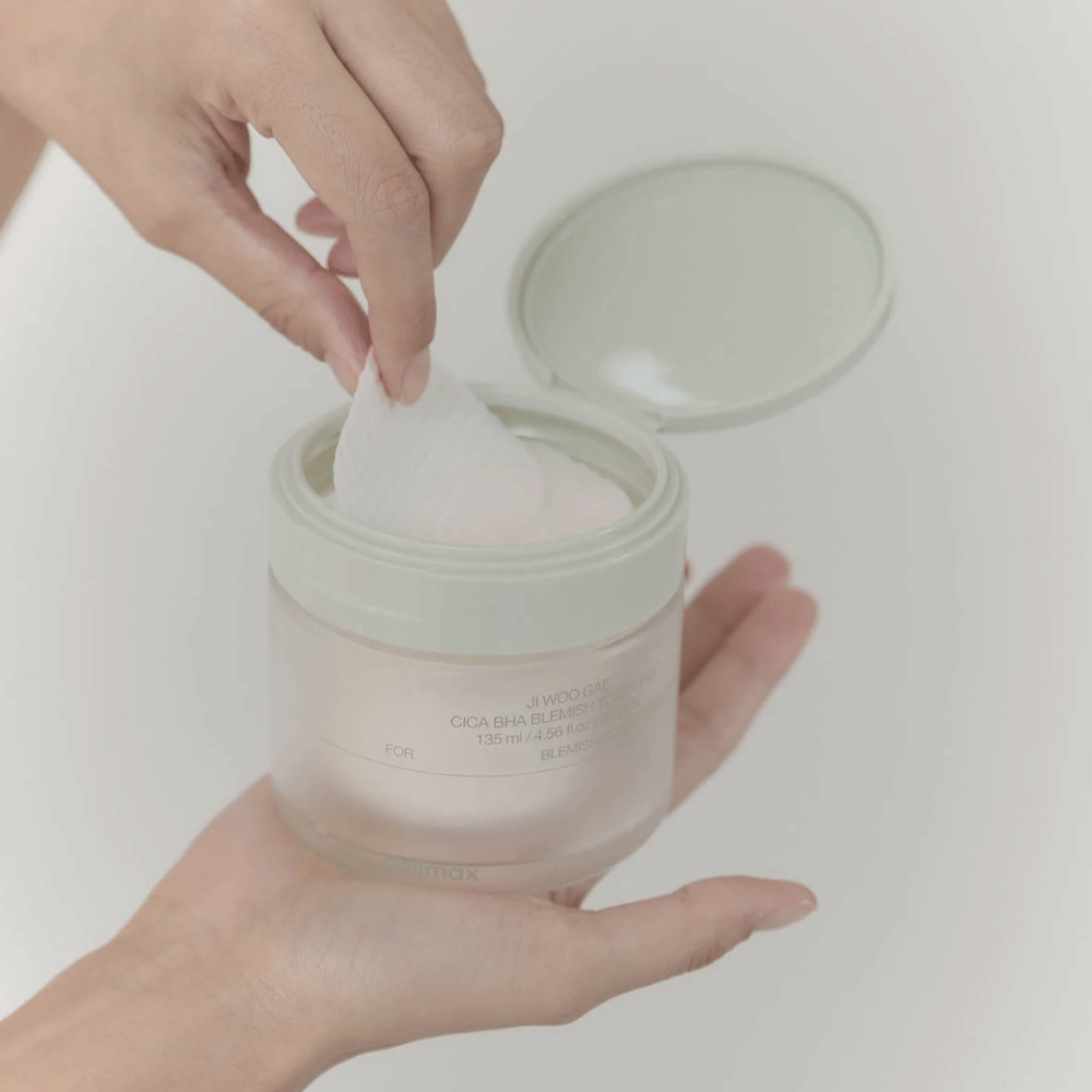 Une main prend un tampon tonifiant du pot Celimax Ji Woo Gae Cica BHA Blemish Toner Pad, parfait pour les peaux impures.