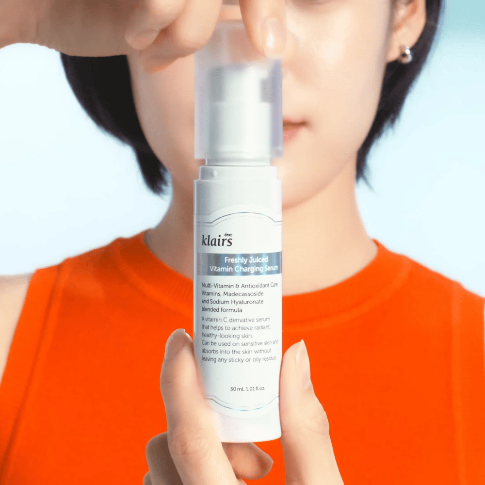 Femme en pull orange tenant Klairs Freshly Juiced Vitamin Charging Serum devant elle sur fond bleu.