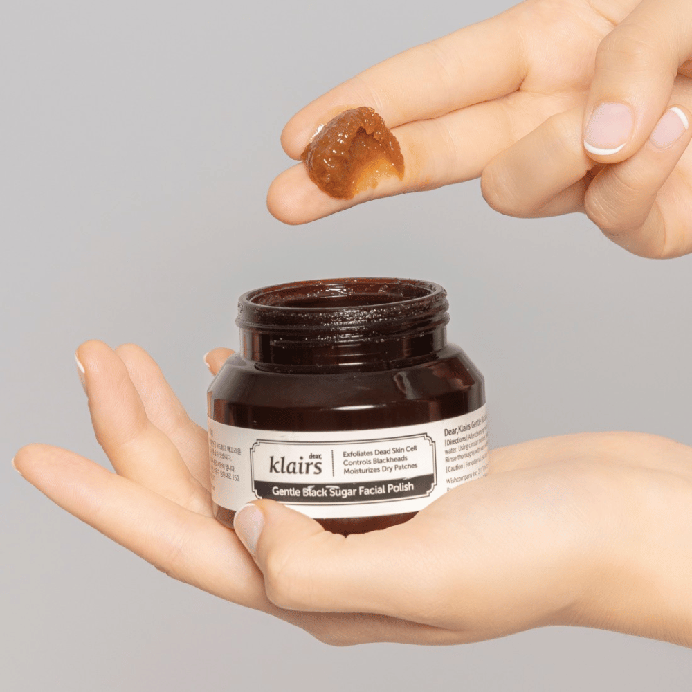 Des mains tiennent un pot ouvert de Klairs Gentle Black Sugar Facial Polish et montrent le produit sur un doigt, sur un fond gris.