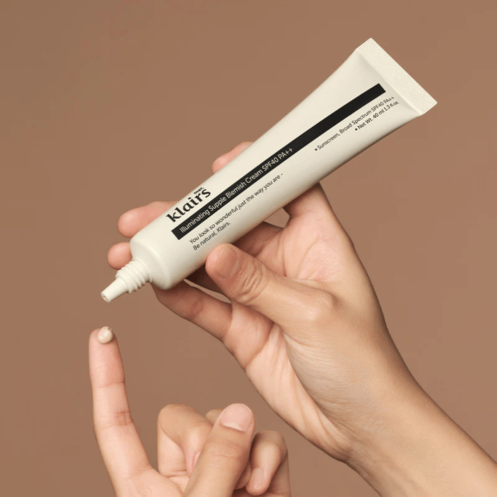 Une main tient un tube de Klairs Illuminating Supple Blemish Cream SPF40 PA++ et presse une petite quantité de crème sur un doigt, sur un fond brun.