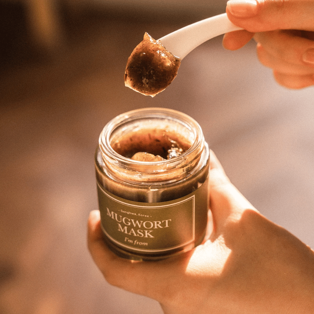Gros plan d'une main tenant I'm From Mugwort Mask tandis qu'une cuillère soulève le masque gel à base de plantes du pot, dans une lumière naturelle chaude.