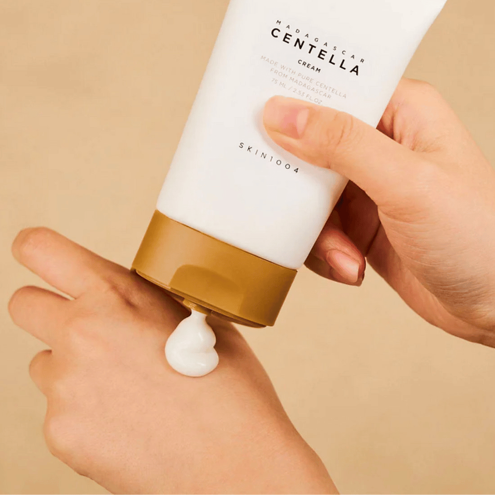 L'image montre une personne tenant un tube de "Madagascar Centella Cream" de SKIN1004. C'est une crème pour la peau fabriquée à partir de Centella pure de Madagascar, de taille 75 ml / 2,53 fl.oz. La crème est appliquée sur la main et est destinée à nourrir et hydrater la peau.