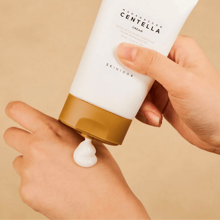 L'image montre une personne tenant un tube de "Madagascar Centella Cream" de SKIN1004. C'est une crème pour la peau fabriquée à partir de Centella pure de Madagascar, de taille 75 ml / 2,53 fl.oz. La crème est appliquée sur la main et est destinée à nourrir et hydrater la peau.