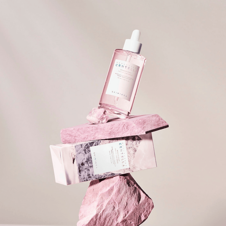 Flacon transparent avec ampoule de Centella rose de SKIN1004, équilibré sur des blocs de pierre, avec un emballage en carton en dessous, sur un fond neutre.