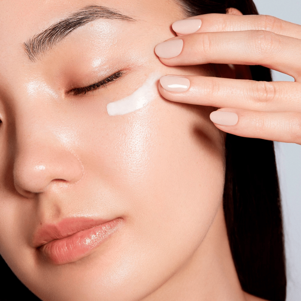 Une femme applique délicatement une crème pour les yeux blanche sous l'œil. Sa peau semble radieuse et hydratée, ce qui souligne l'effet de la crème pour les yeux. Cela met en avant le produit comme étant à la fois hydratant et adapté à la peau sensible autour des yeux.