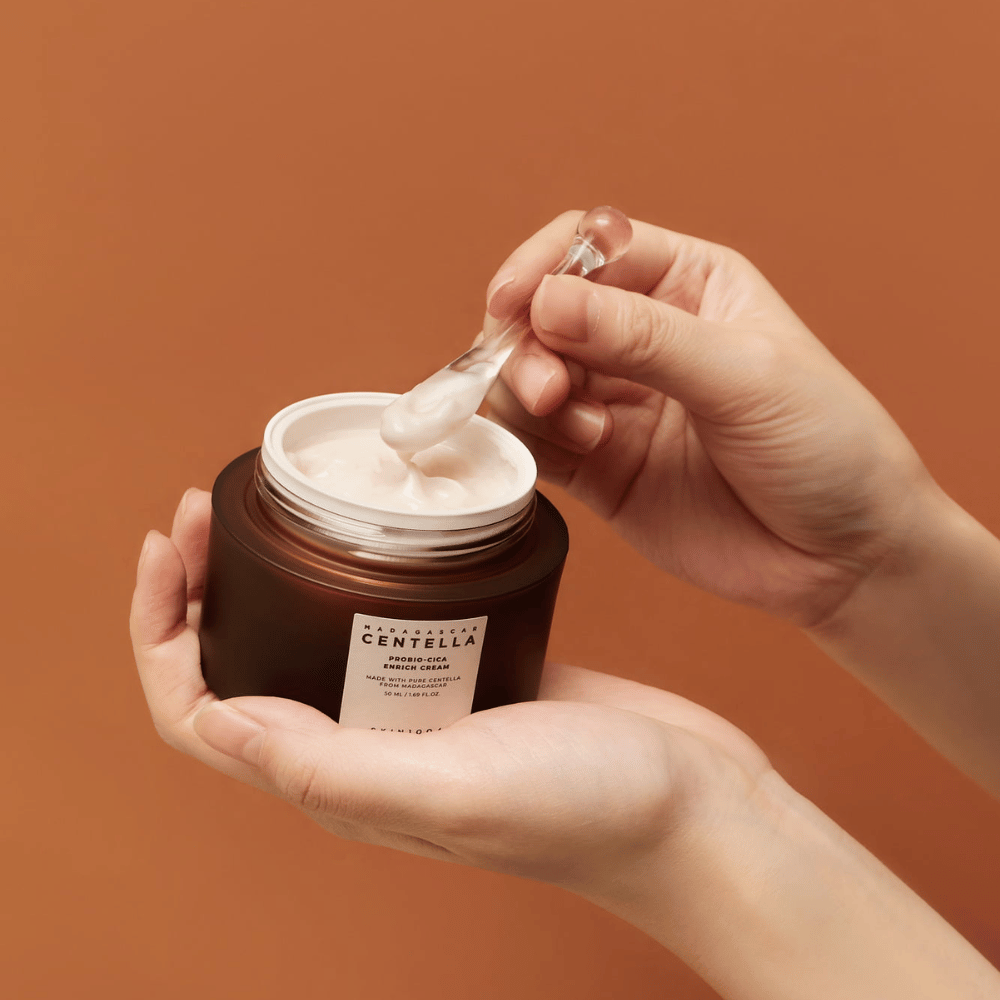 Mains tenant un pot ouvert de Madagascar Centella Probio-Cica Enrich Cream et une spatule avec de la crème sur un fond de couleur terracotta uniforme.