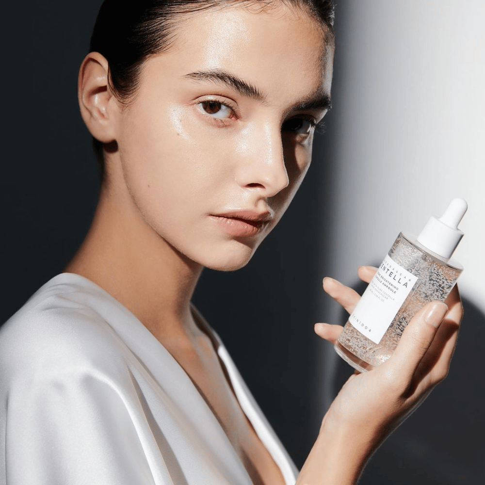 Sur l'image, on voit une personne tenant une bouteille avec un applicateur compte-gouttes. La bouteille est étiquetée "SKIN1004 Madagascar Centella Tone Brightening Capsule Ampoule". La personne a un teint chatoyant qui reflète la lumière, ce qui suggère un effet possible du produit.