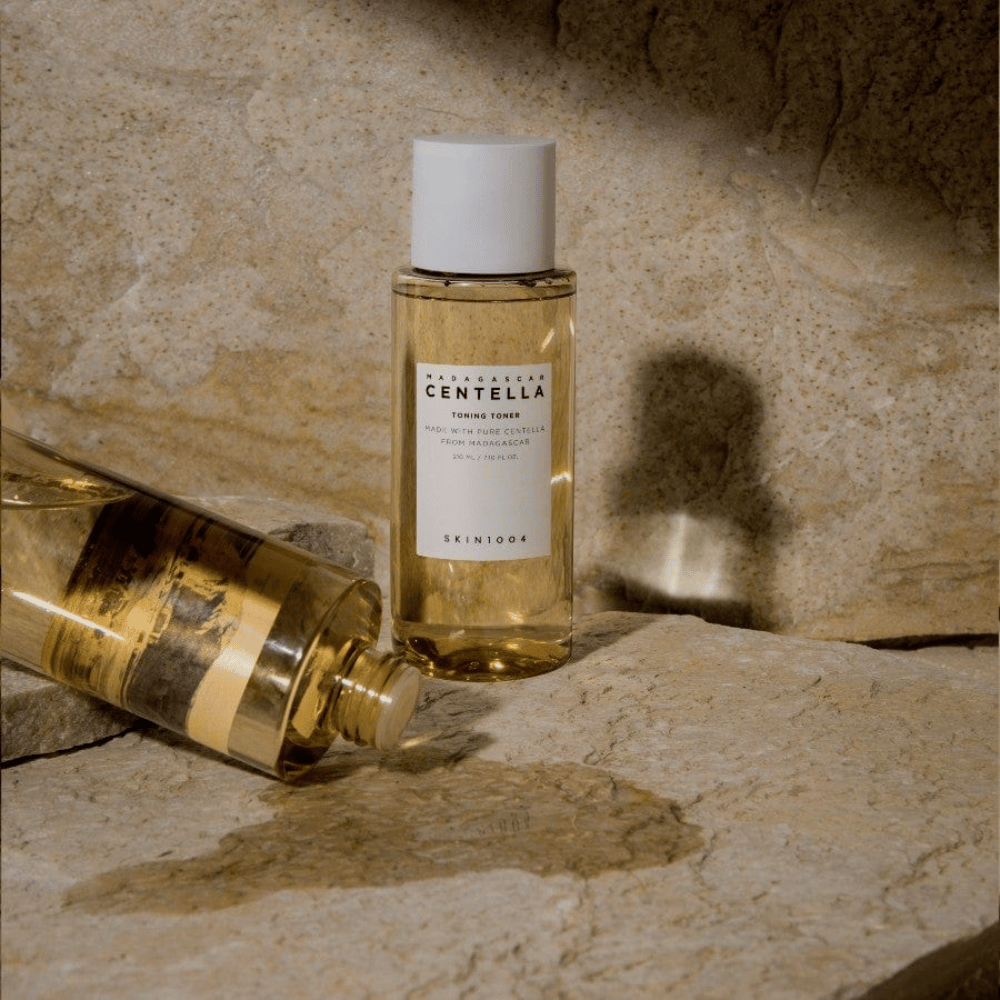 L'image montre une bouteille "Madagascar Centella Toning Toner" de SKIN1004. C'est une bouteille transparente remplie d'un liquide jaunâtre, probablement un tonique pour la peau. Il est indiqué qu'elle est faite avec de la centella pure de Madagascar et que le contenu est de 210 ml / 7,10 fl.oz. La bouteille et une bouteille couchée sont placées sur une surface rocheuse, ce qui donne une impression naturelle et minimaliste.