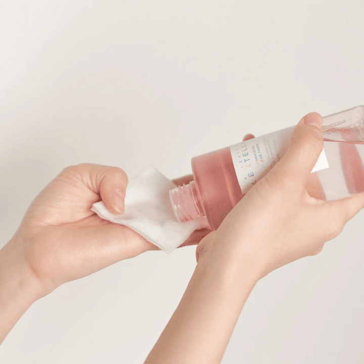 Une personne verse Madagascar Centella Poremizing Clear Toner d'une bouteille transparente sur un coton. Le produit, qui a une teinte rose, est utilisé pour rafraîchir et nettoyer la peau. Les mains sont visibles, montrant l'application du toner.