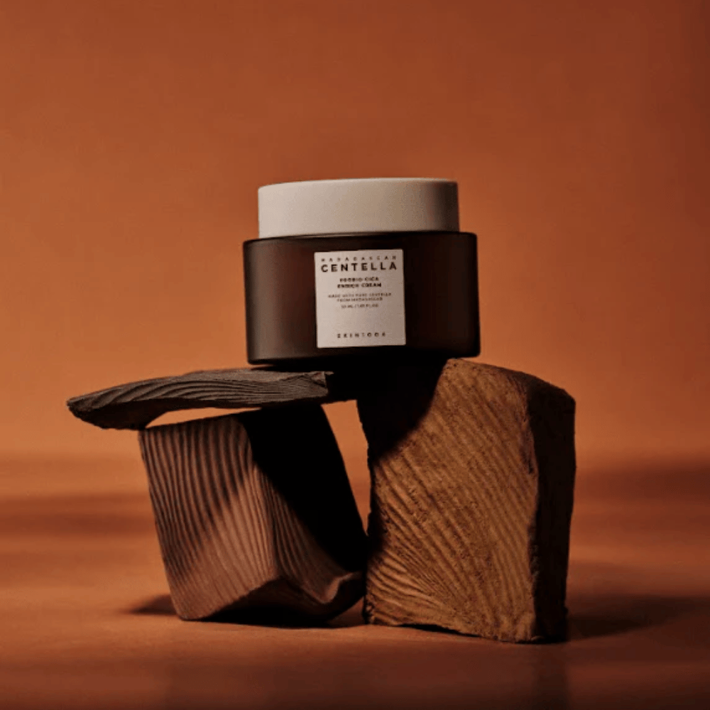 SKIN1004 Madagascar Centella Probio-Cica Enrich Cream dans un pot en verre brun élégant sur des blocs de bois décoratifs – crème pour le visage coréenne nourrissante qui renforce la barrière cutanée.