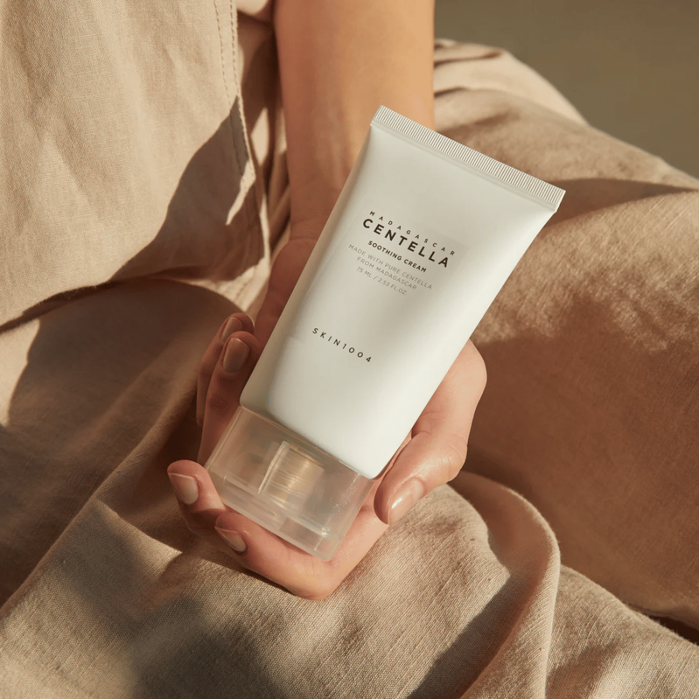 Une personne tient un tube blanc de Madagascar Centella Soothing Cream de SKIN1004. Le tube a un bouchon transparent et un texte décrivant le produit comme apaisant et fabriqué avec de la Centella pure. La personne est habillée de vêtements clairs.