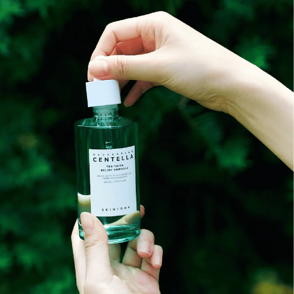 Personne ouvre SKIN1004 Madagascar Centella Tea-Trica Relief Ampoule – bouteille en verre vert avec étiquette blanche tenue dans les mains devant un fond verdoyant.