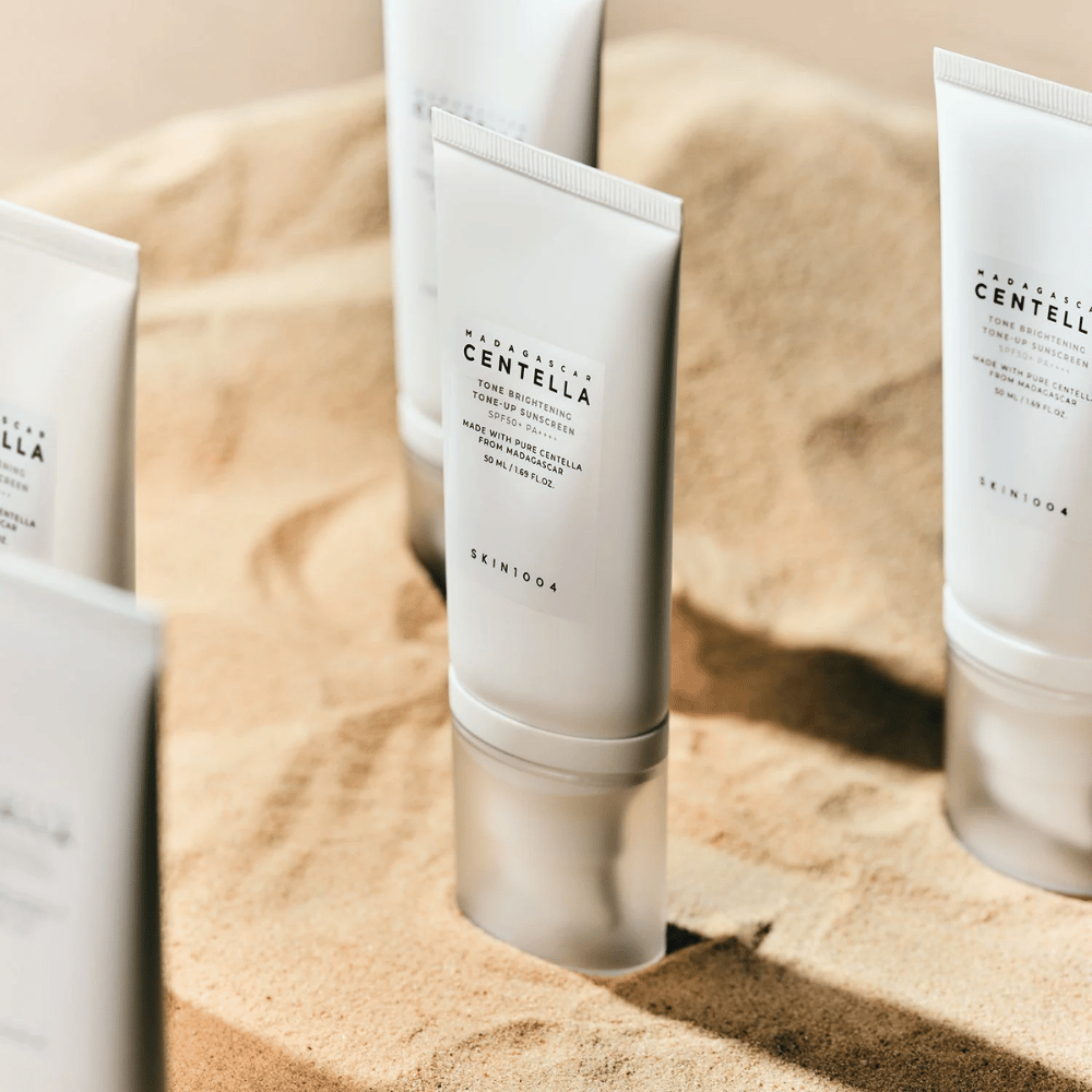 Plusieurs tubes avec Madagascar Centella Tone Brightening Tone-Up Sunscreen se tiennent dans le sable. Le tube est blanc avec du texte noir et contient 50 ml. Le produit a SPF 50+ PA++++ et est fabriqué avec de la Centella pure de Madagascar.