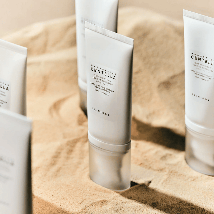 Plusieurs tubes avec Madagascar Centella Tone Brightening Tone-Up Sunscreen se tiennent dans le sable. Le tube est blanc avec du texte noir et contient 50 ml. Le produit a SPF 50+ PA++++ et est fabriqué avec de la Centella pure de Madagascar.