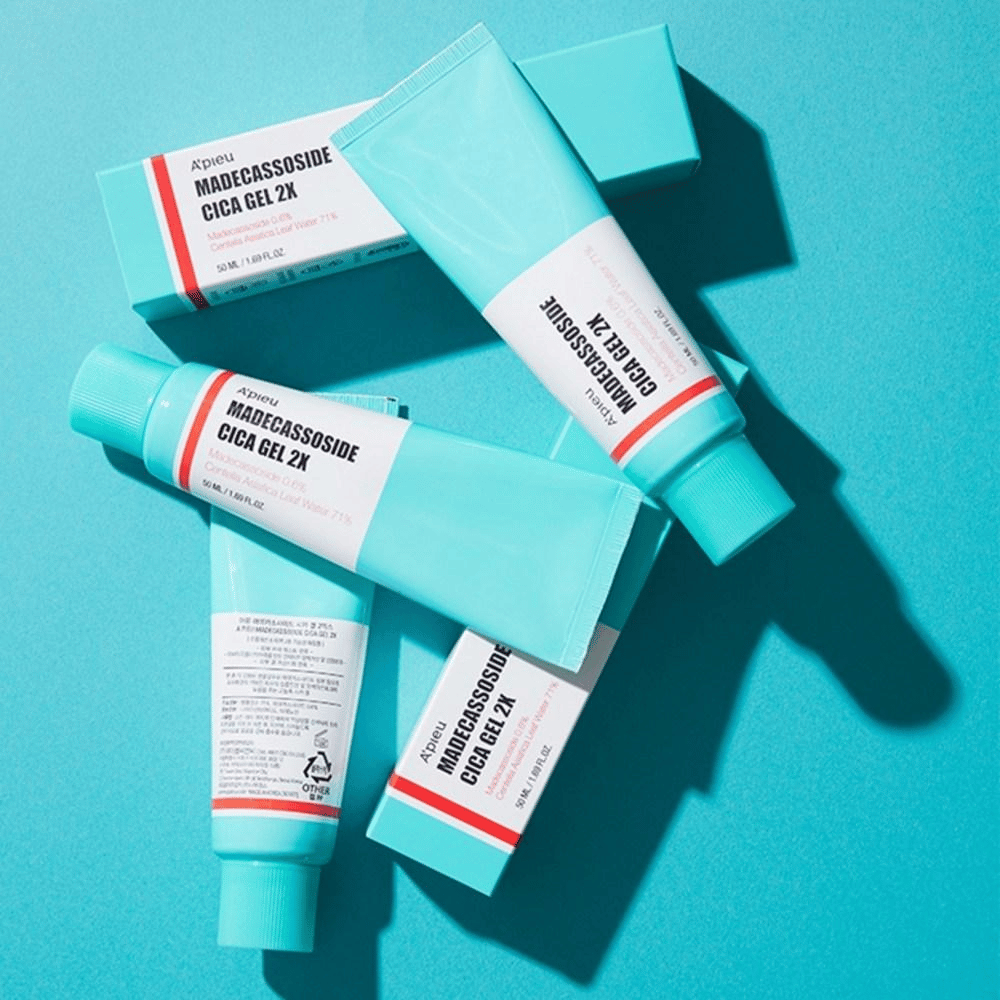 Plusieurs tubes et cartons de A’pieu Madecassoside Cica Gel 2X en emballage turquoise avec étiquette blanche et rouge, posés sur un fond turquoise sous une lumière vive.