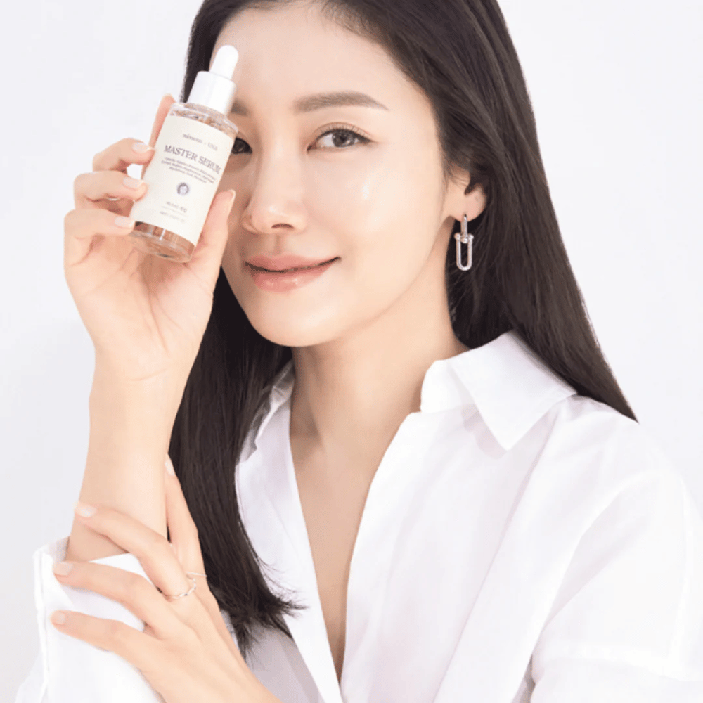 Femme souriante avec de longs cheveux foncés et une chemise blanche tenant Mixsoon Master Serum à côté de son visage. Sa peau semble éclatante et hydratée.