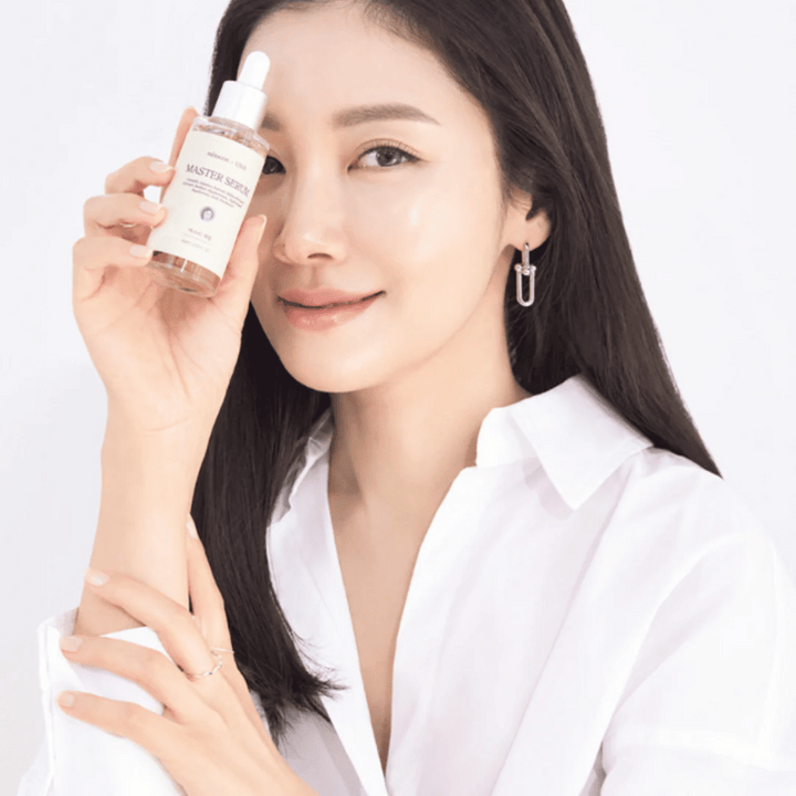 Femme souriante avec de longs cheveux foncés et une chemise blanche tenant Mixsoon Master Serum à côté de son visage. Sa peau semble éclatante et hydratée.