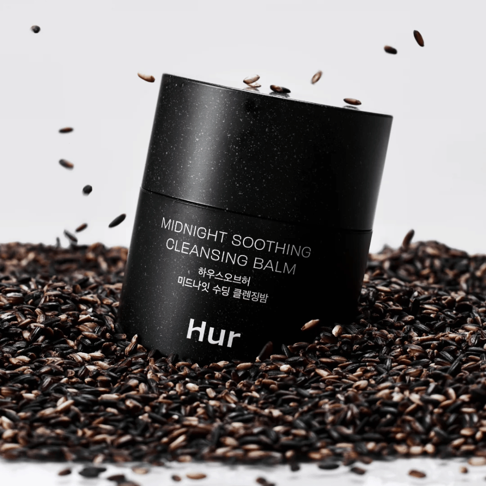Comment Midnight Soothing Cleansing Balm dans un pot noir placé sur des graines noires. Baume nettoyant apaisant qui dissout le maquillage et les impuretés tout en prenant soin de la peau.