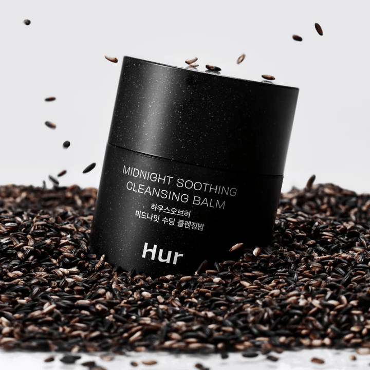 Comment Midnight Soothing Cleansing Balm dans un pot noir placé sur des graines noires. Baume nettoyant apaisant qui dissout le maquillage et les impuretés tout en prenant soin de la peau.
