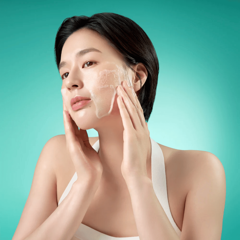 Une femme nettoie son visage avec une mousse nettoyante douce sur un fond vert.