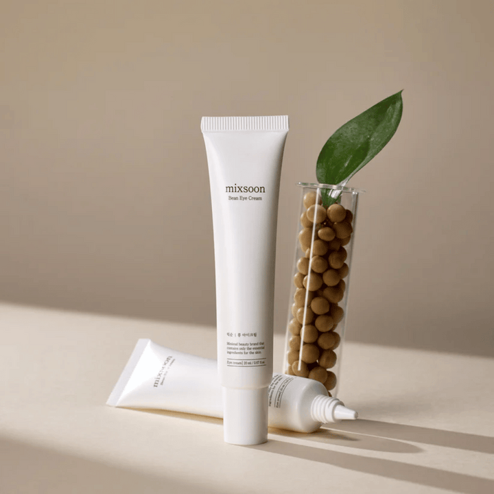 Deux tubes Mixsoon Bean Eye Cream placés à côté d'un tube à essai rempli de graines de soja et d'une feuille verte, sur une surface beige avec des jeux d'ombres.