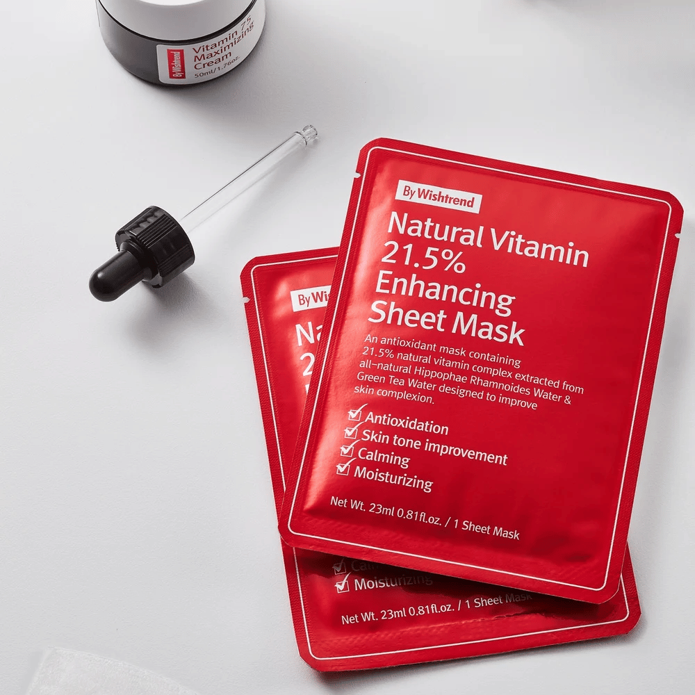 By Wishtrend Natural Vitamin 21.5% Enhancing Masque tissu dans des emballages rouges, placés sur une surface blanche avec une pipette et un pot de crème pour le visage.