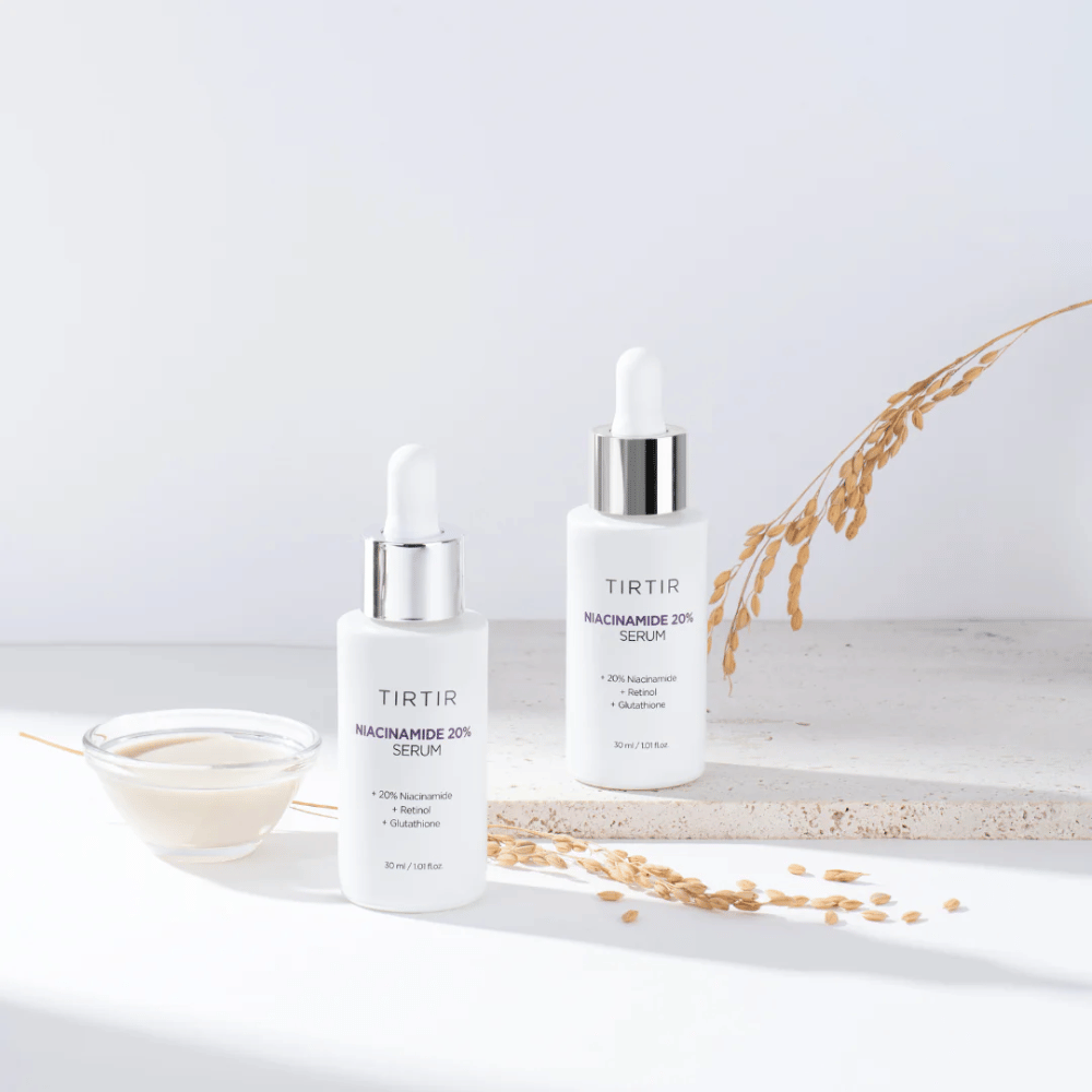 Deux bouteilles de Tirtirs "Niacinamide 20% Serum" sont présentées sur une surface claire avec un petit bol contenant une substance crémeuse et quelques brins de plantes sèches. Le sérum contient 20 % de niacinamide, rétinol et glutathion, ce qui contribue à améliorer l'apparence et la texture de la peau.