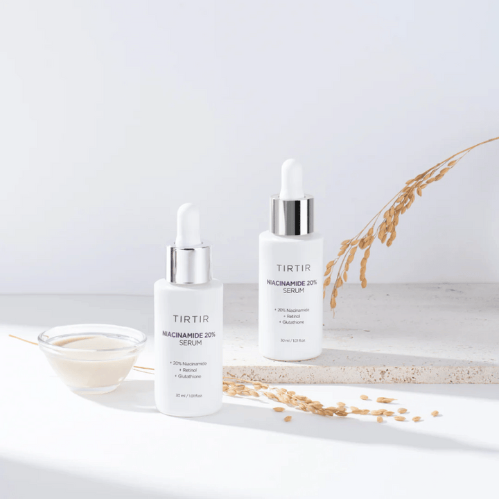 Deux bouteilles de Tirtirs "Niacinamide 20% Serum" sont présentées sur une surface claire avec un petit bol contenant une substance crémeuse et quelques brins de plantes sèches. Le sérum contient 20 % de niacinamide, rétinol et glutathion, ce qui contribue à améliorer l'apparence et la texture de la peau.