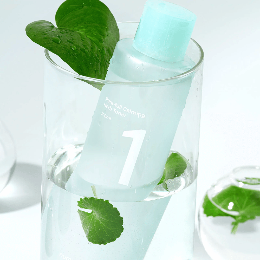 Une bouteille avec No.1 Pure-Full Calming Herb Toner placée dans un verre avec de l'eau et des feuilles vertes. La bouteille a un bouchon bleu clair et une étiquette avec le texte "Pure-full Calming Herb Toner" ainsi que "300ml". Le produit a l'air frais avec de l'eau et de la végétation en arrière-plan, ce qui donne une sensation apaisante et naturelle.
