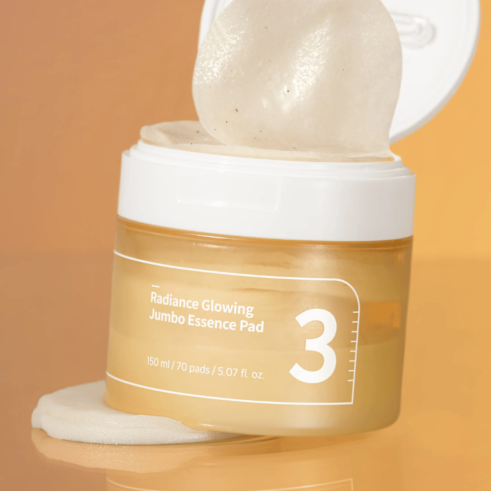Pot ouvert avec numbuzin No. 3 Radiance Glowing Jumbo Essence Pad, un tampon visible incliné contre le bord, fond en ton orange.