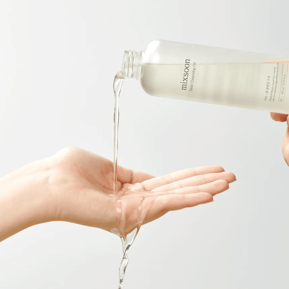 Une huile nettoyante claire est versée d'une bouteille transparente avec une étiquette blanche où il est écrit "MixSoon" dans une paume ouverte. L'huile forme un jet brillant entre l'ouverture de la bouteille et la main, démontrant la consistance fluide de l'huile. La main est immobile et tenue de manière à recueillir toute la quantité d'huile nettoyante sur un fond uni et clair.
