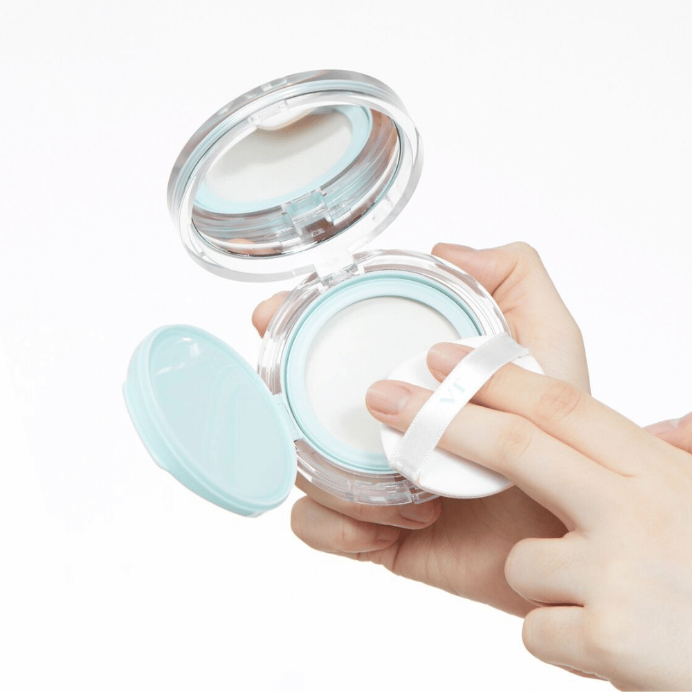 Une main tient un pot ouvert avec VT PDRN Essence Glow Sun Pact. Le tampon est sur le point d'appliquer la poudre, qui a une surface blanche et un bord bleu autour du pot.