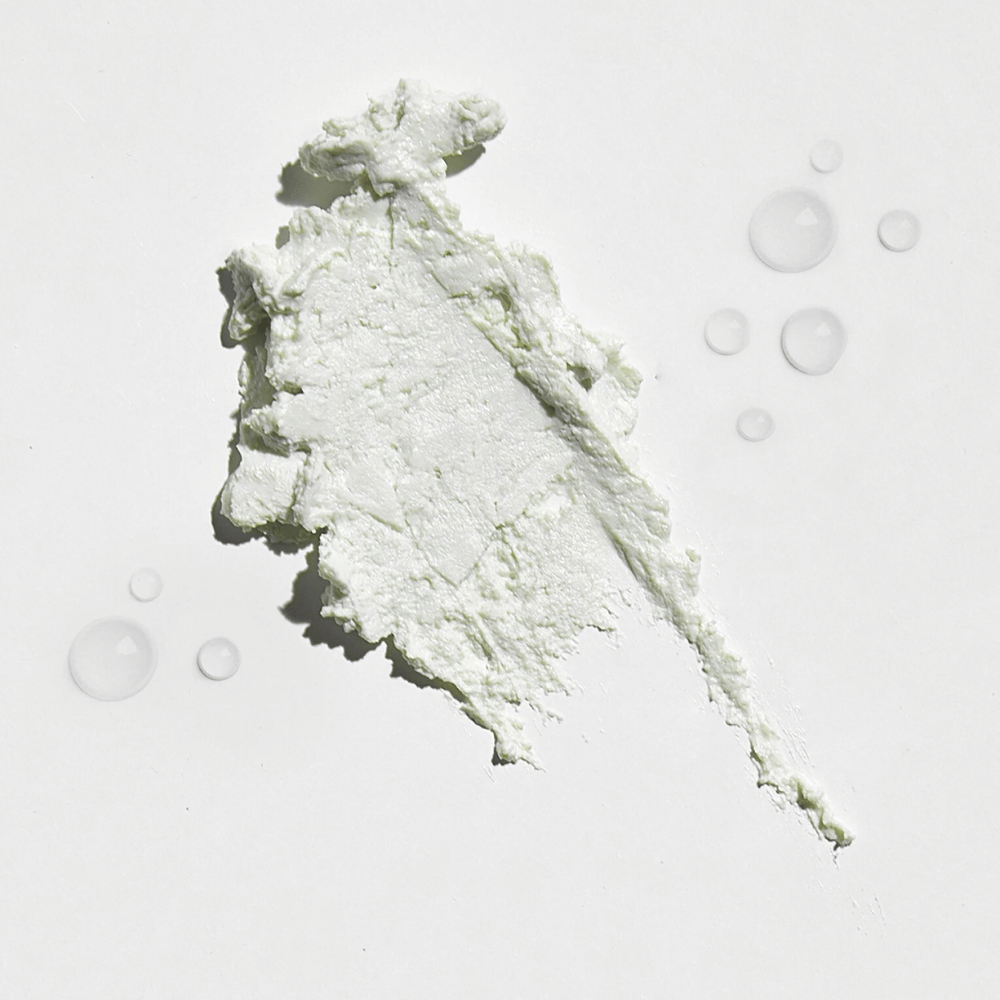 Gros plan sur une substance crémeuse blanche sur un fond blanc, avec quelques gouttes d'eau autour. La texture semble être une poudre solaire ou foundation avec une légère lueur.