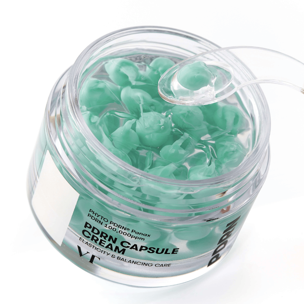 Gros plan sur un pot ouvert de VT PDRN Capsule Cream. Le contenu se compose de petites capsules vertes dans un gel transparent, avec une spatule fournie pour l'application. Le texte sur le pot indique "Elasticity & Balancing Care".
